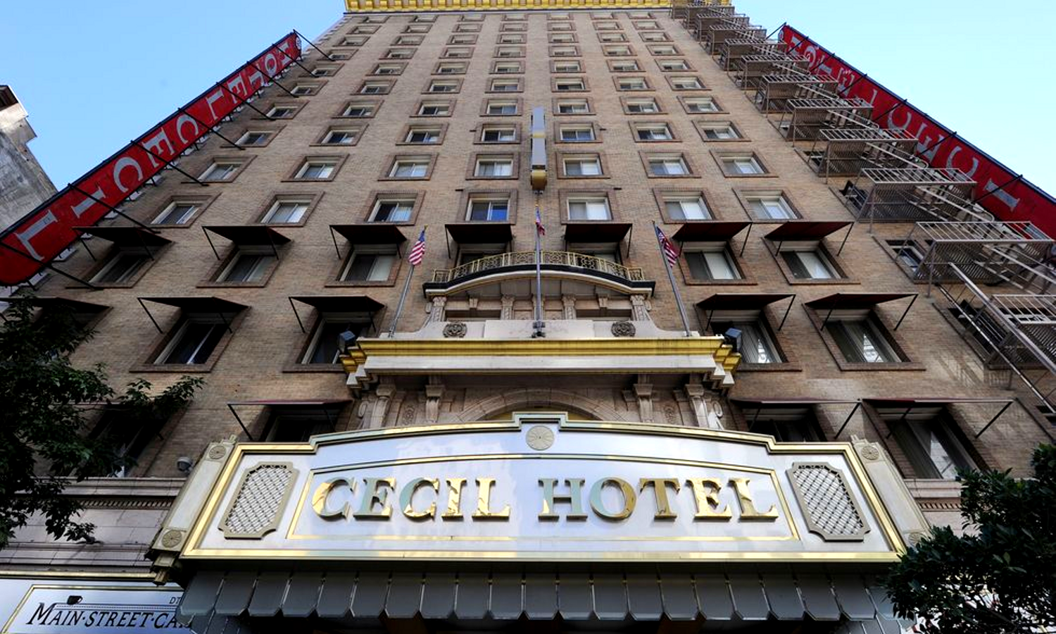 Picture of large multi-story building. The name 'Cecil Hotel' is on gold font in the front and American flags are mounted on the building. 