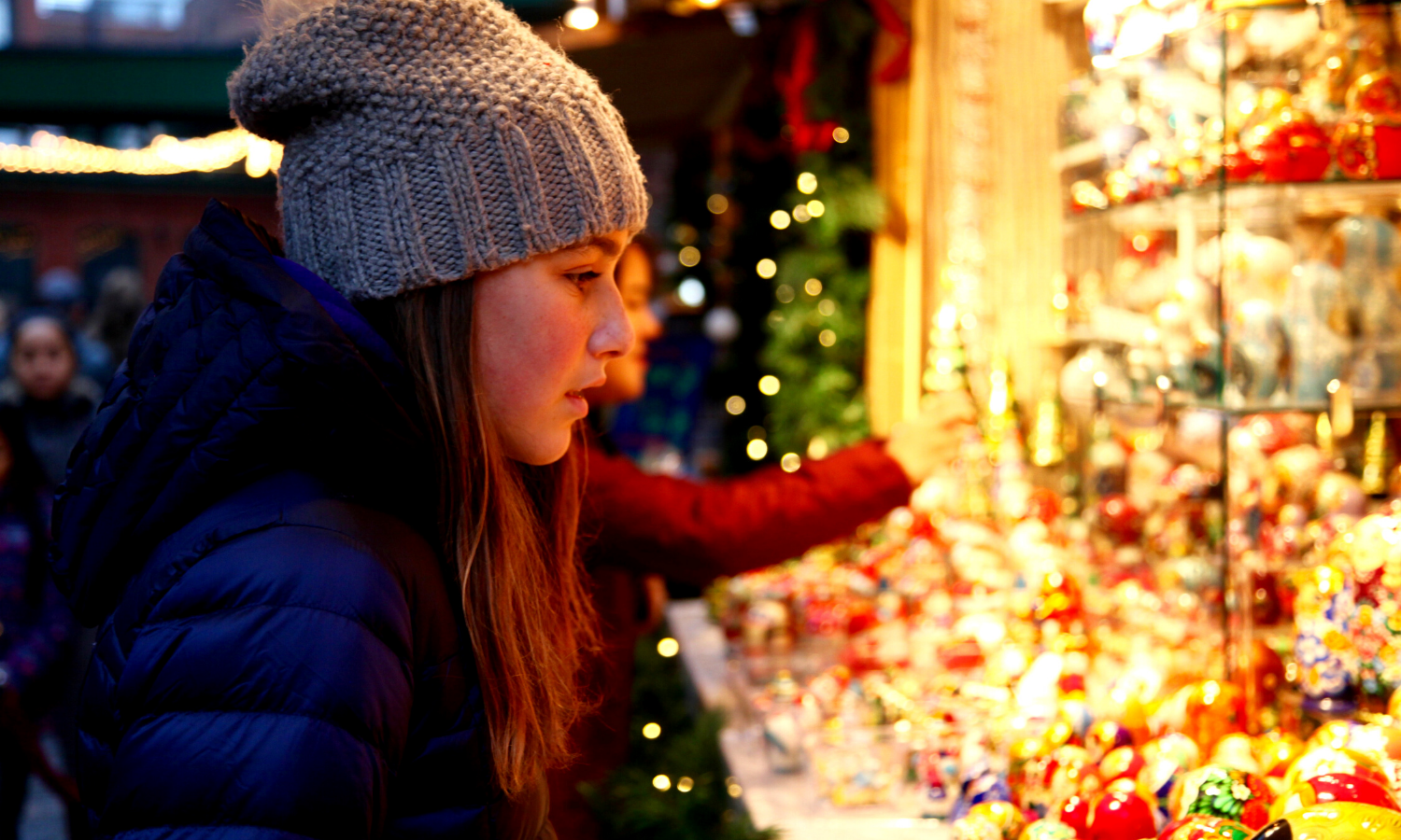 Picture of a teenage girl in a hat looking a variety of crafts in a stall. There are festive fairy lights and Christmas trees in the background. 