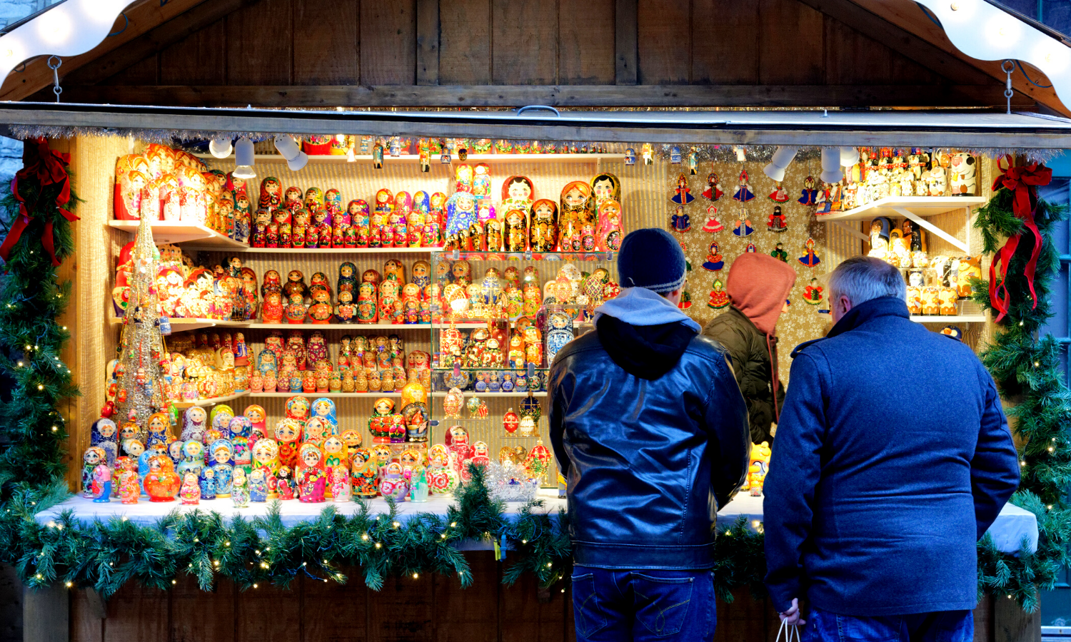 A photograph of two men looking at a stall showcasing handmade Russian Dolls. There are fairy lights and fir tinsel surrounding the hut. 