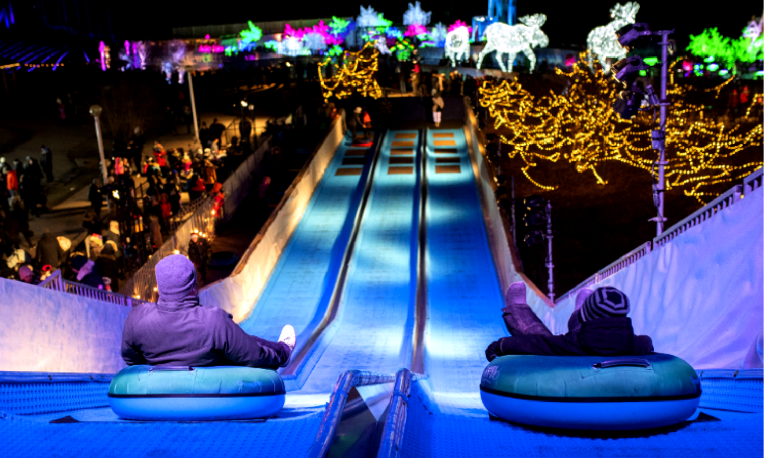 A picture of two people going down a slide in inflatable doughnuts. There are festive lights in the background. 