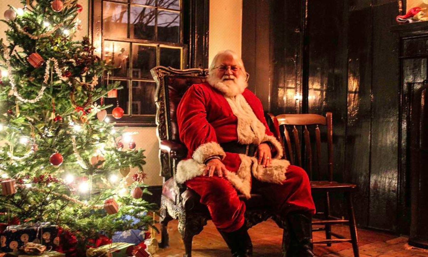 A picture of Santa Claus sat in a huge armchair next to a Christmas tree. 
